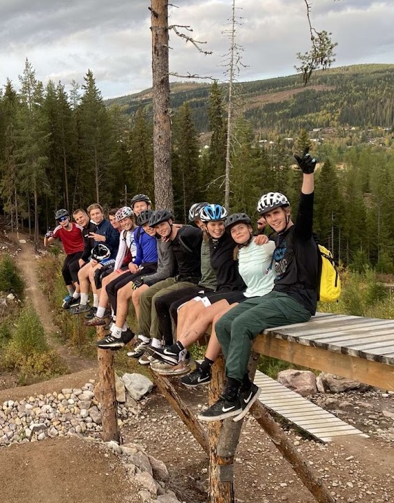 En rekke med ungdommer sitter på en trebenk langt over bakken. På hodet har de hjelmer. Noen går i shorts men andre går i bukse. Tørt landskap.