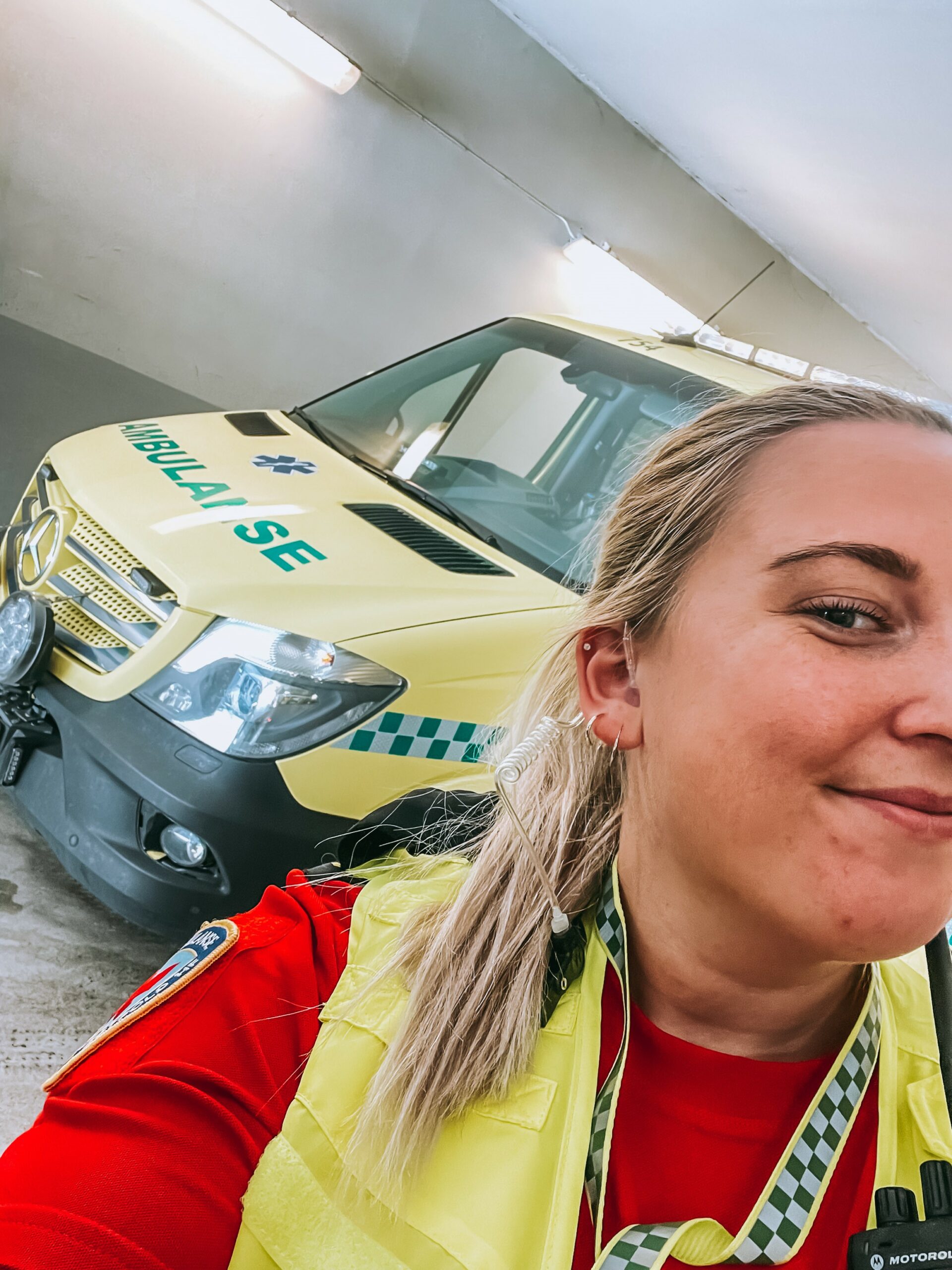 en ung kvinne i ambulanseuniform som tar selfie med en ambulanse i bakgrunnen