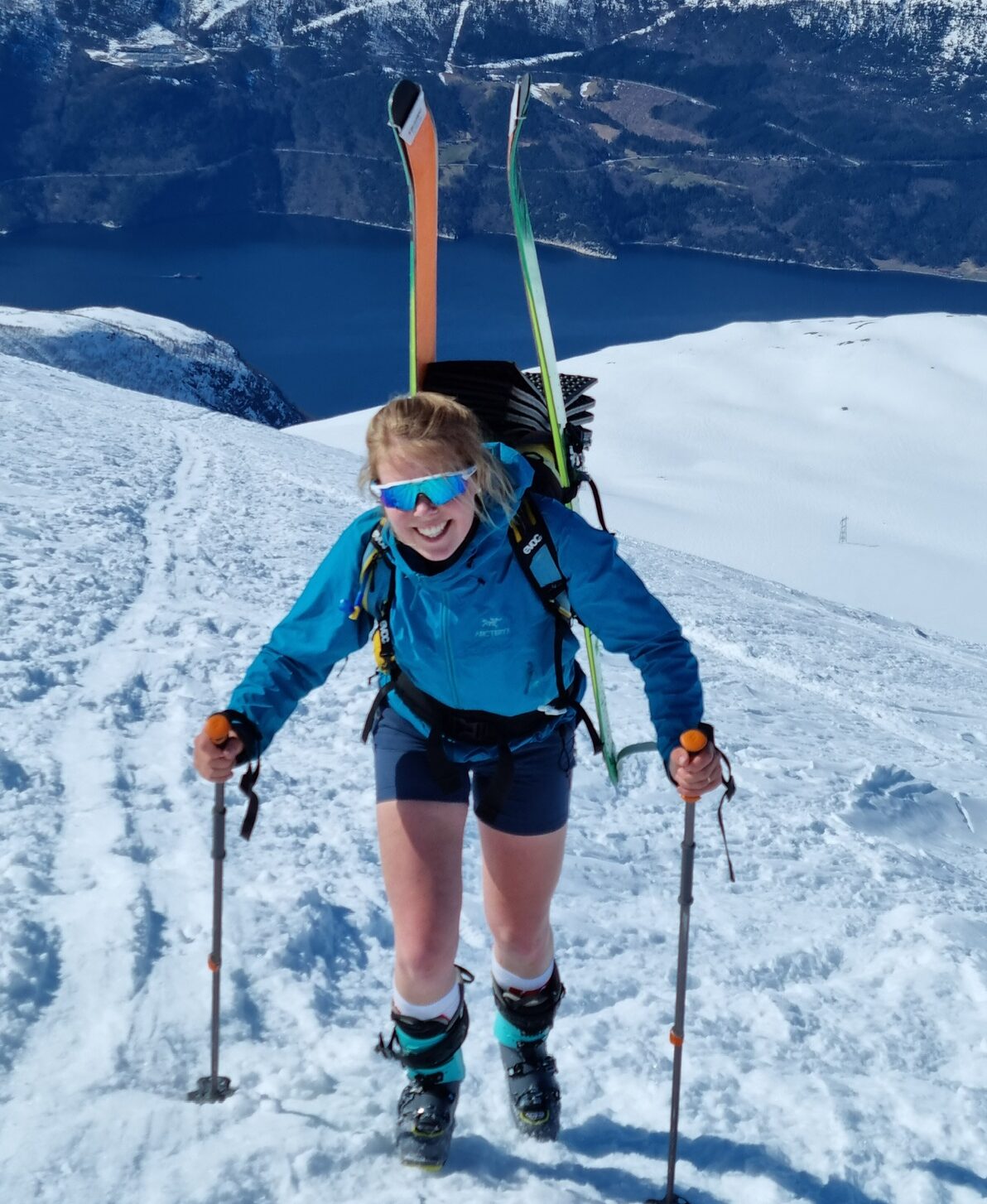 en ung kvinne går oppover et snødekt fjell med ski på ryggen og shorts