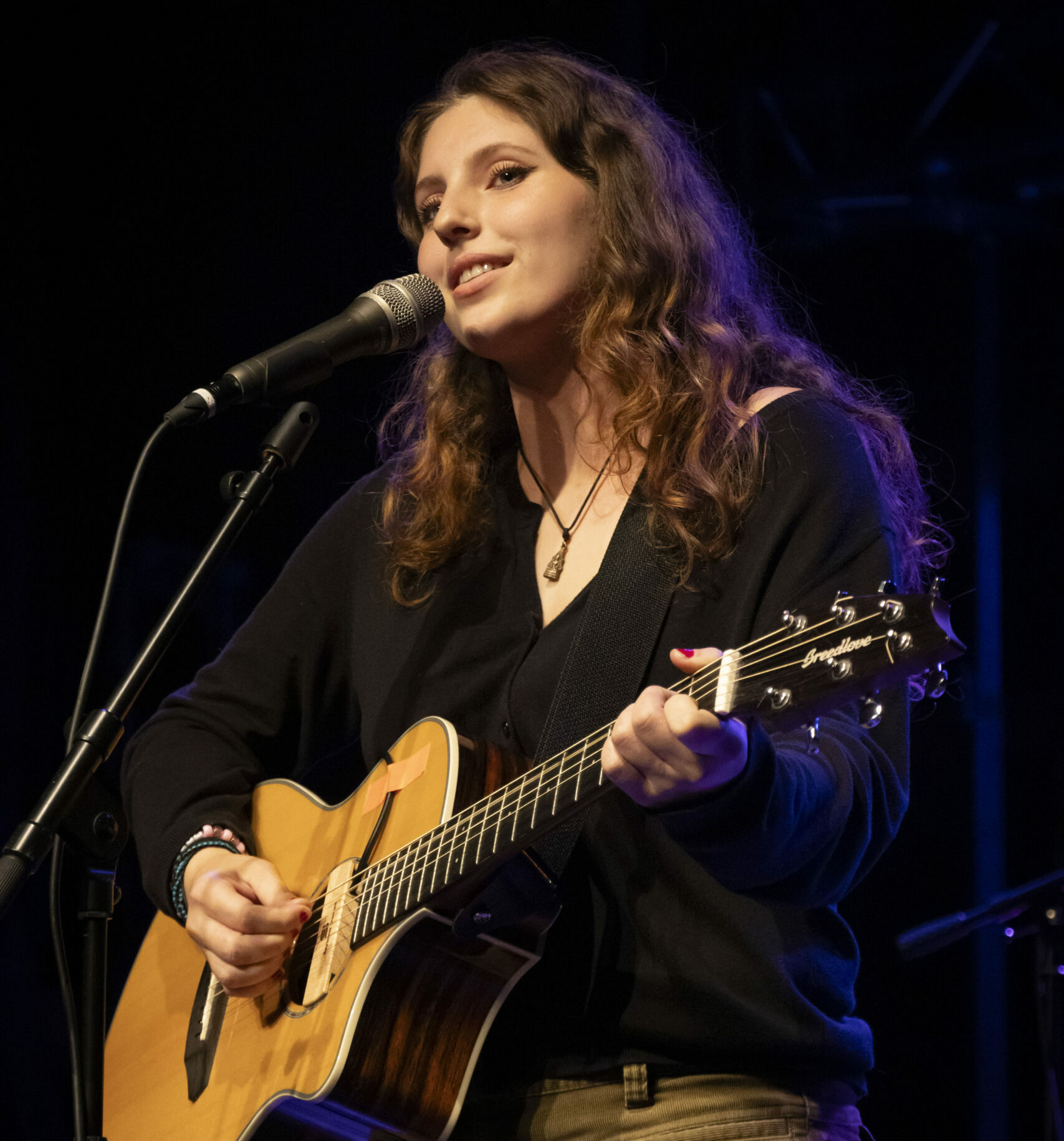 En person med langt hår står med gitar og synger inn i mikrofon. 