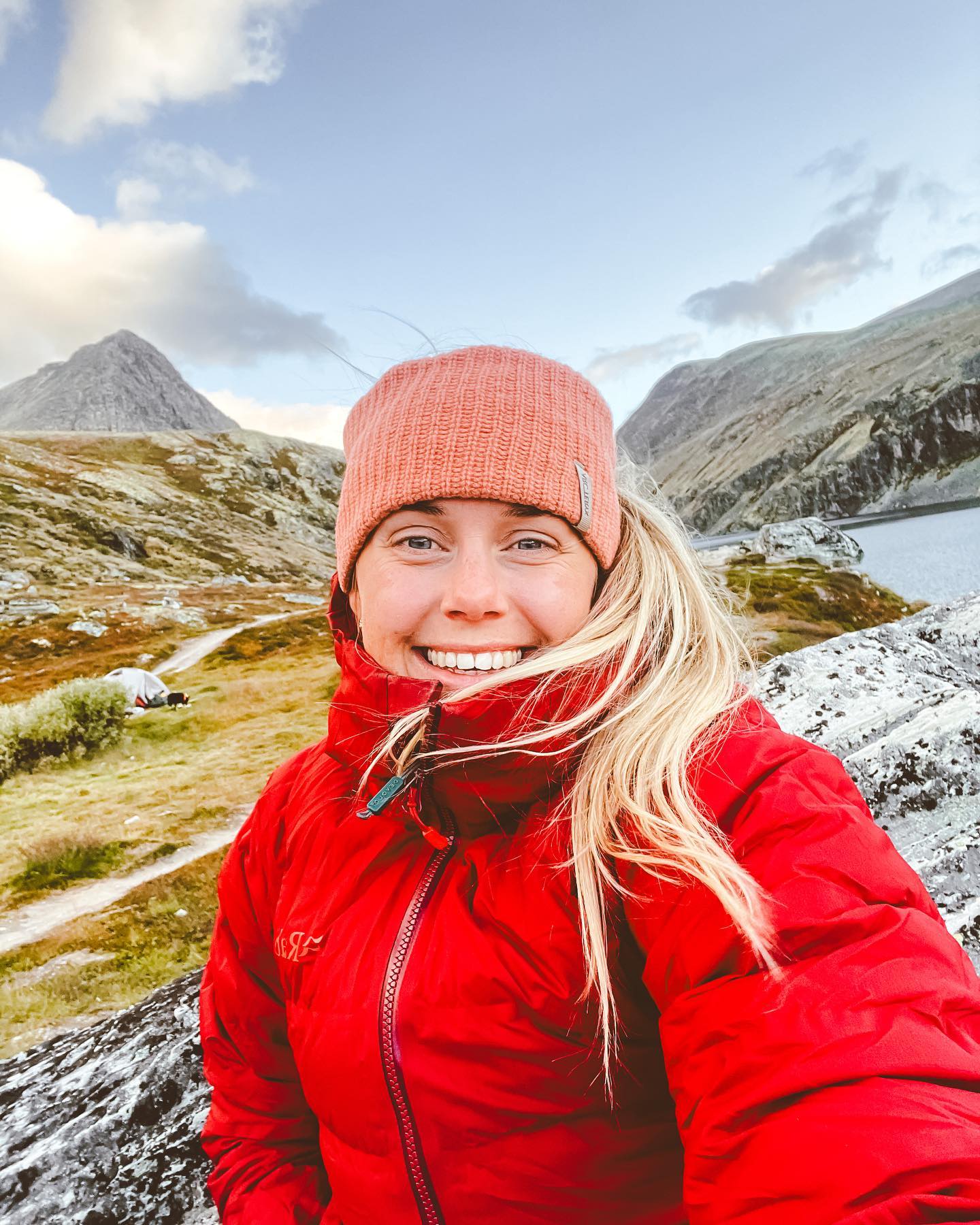 En lyshåret jente smiler i kamera med rød jakke. 